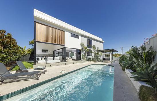 Coolum Beach Prize Home showcasing the pool area with a cabana and deck chairs surrounded by lush gardens. The covered alfresco area is visible behind the pool.