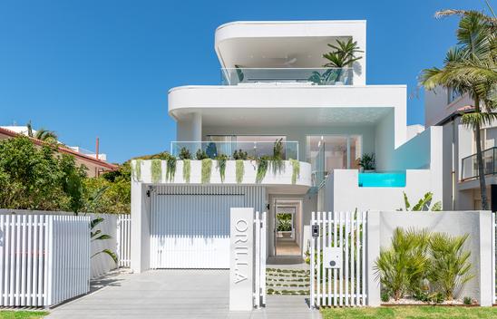 External image of a new three-level tiered home. Second floor has a balcony with plants. First floor has a raised pool to the right, and a covered entertaining area to the left, with plants spilling over the edge. Ground floor shows a closed garage, open front gate and front door with a clear view into the house to see the back courtyard. The name of the house on the front is Orilla.