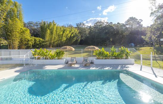 A pool with two lounge chairs with umbrellas and beds of garden either side. A large backyard is in the background.