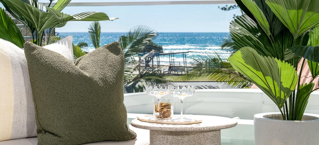 Second floor covered entertainment area with views to the beach. Cropped shot showing a clear view to the beach, a corner of a white couch with a green cushion and a round marble table with two glasses. Potted palms to either side.