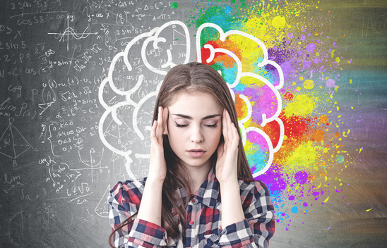 young woman pressing hands against temples with her eyes closed in front of a backboard with a brain drawing, colours splashing out from one side