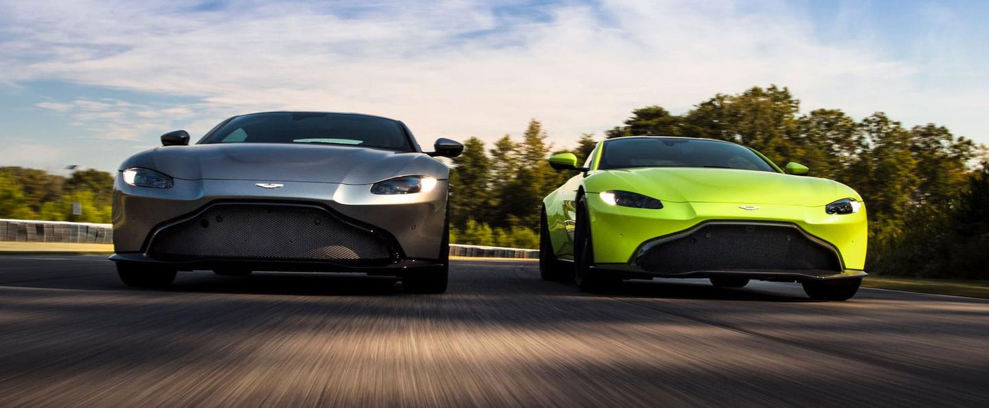 Image of two Aston Martin Vantages driving side by side. The car on the left is a dark grey colour and the car on the right is a lime green colour. Both are facing the camera and seem to be driving at high speed. The image is taken low from the ground.