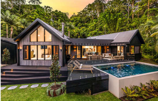 yourtown Prize Home Draw 545. Dusk external image from the front of a single-level pavilion-style house with black cladding. There is an alfresco area at the front with a pool. A garage and driveway is on the left hand side. Lush gardens surround the home.