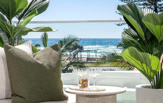 Second floor covered entertainment area with views to the beach. Cropped shot showing a clear view to the beach, a corner of a white couch with a green cushion and a round marble table with two glasses. Potted palms to either side.